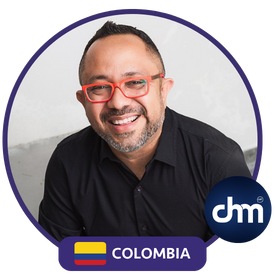 Fotografía de Raúl Alfonso Camacho Rodríguez sonriendo, con gafas rojas y camisa negra, acompañado del logotipo de DMI Colombia sobre un fondo claro.