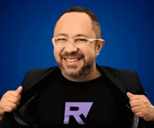 Fotografía de Raúl Alfonso Camacho Rodríguez sonriendo frente a un fondo azul, vistiendo gafas y camiseta negra con una “R” morada, acompañado de saco negro.