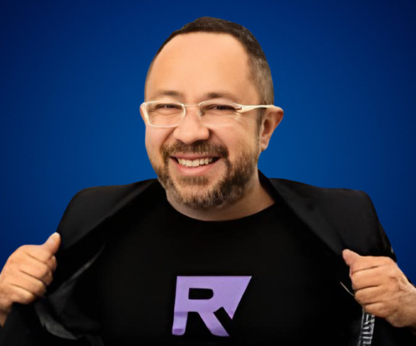 Fotografía de Raúl Alfonso Camacho Rodríguez sonriendo frente a un fondo azul, vistiendo gafas y camiseta negra con una “R” morada, acompañado de saco negro.