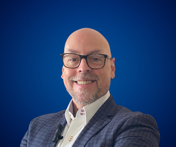 Fotografía de Sal Barrena, sonriendo con traje formal y fondo azul sólido, capturado en un estilo profesional de estudio. Lleva gafas, camisa blanca y micrófono de solapa, proyectando una imagen confiada y cercana.