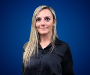 Fotografía de Gabriela Homenuc, de frente, con camisa negra y fondo azul liso, transmitiendo profesionalismo y confianza.