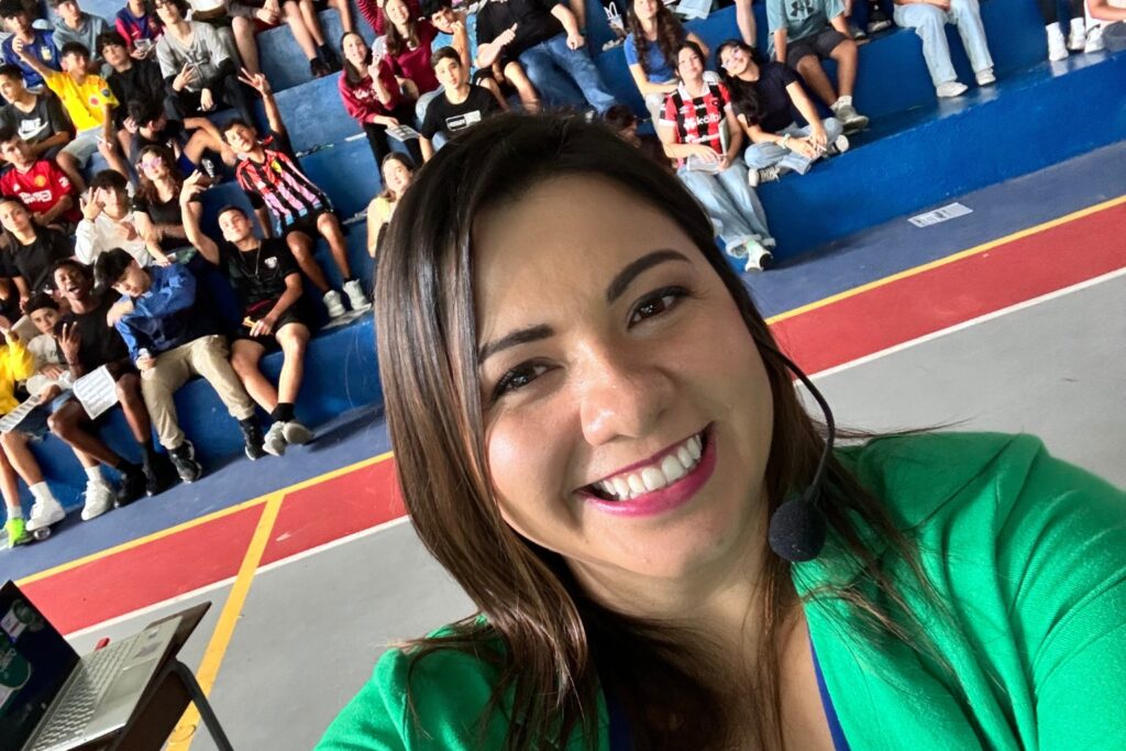 Fotografía de Adriana Castro Bolaños tomando una selfie sonriente con micrófono de diadema, frente a un grupo de jóvenes estudiantes sentados en gradas durante una charla en un gimnasio escolar.