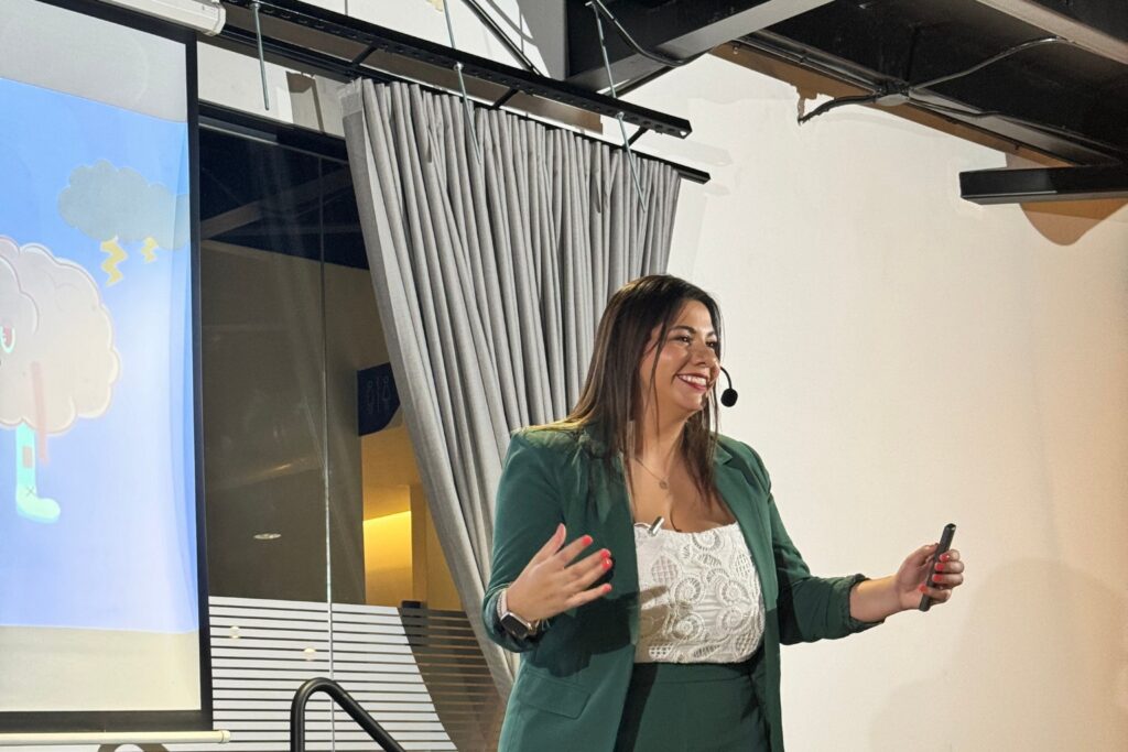 Fotografía de Adriana Castro Bolaños dando una conferencia en interior, sonriendo con micrófono de diadema y control en mano, vestida con traje verde y blusa blanca de encaje, junto a una pantalla con ilustración colorida.