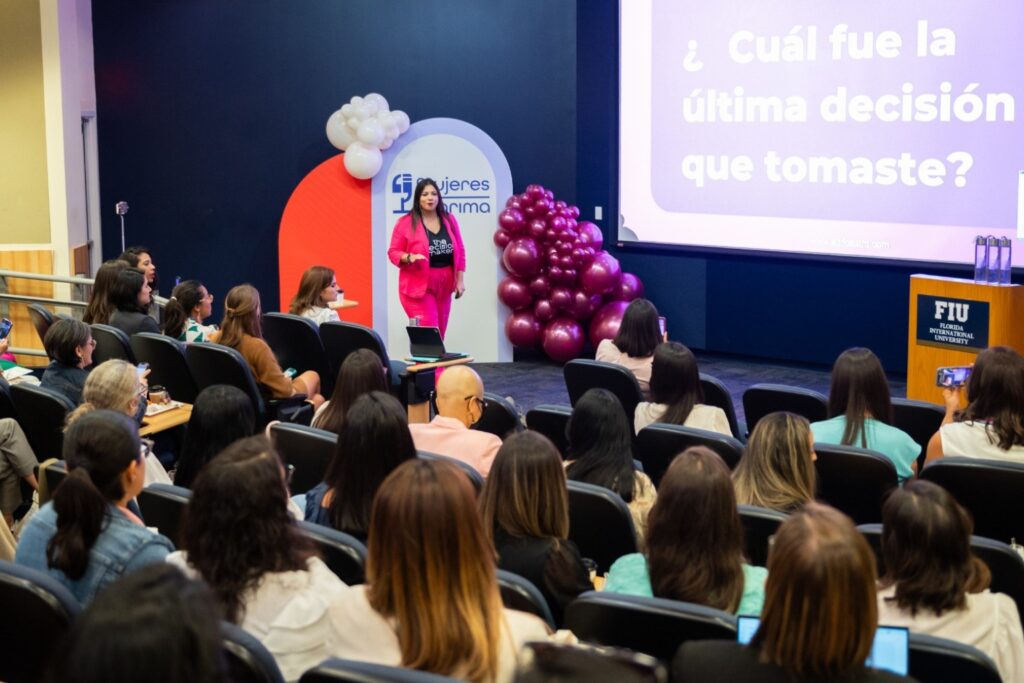 Fotografía de Adriana Castro Bolaños durante una conferencia en Florida International University, vestida con traje fucsia, frente a una audiencia atenta. De fondo, pantalla con la frase “¿Cuál fue la última decisión que tomaste?” y decoración con globos.