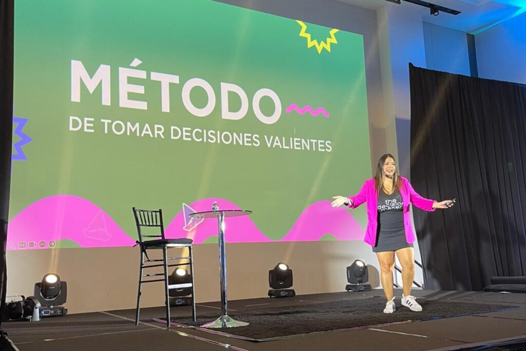 Fotografía de Adriana Castro Bolaños sobre un escenario, con fondo proyectado que dice “Método de tomar decisiones valientes”. Ella viste chaqueta fucsia, vestido negro y zapatillas, en una pose enérgica y sonriente.