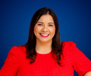 Fotografía de Adriana Castro, sonriendo con una blusa roja vibrante sobre un fondo azul intenso, en un retrato profesional de medio cuerpo.