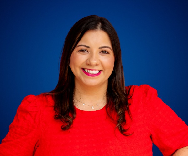 Fotografía de Adriana Castro, sonriendo con una blusa roja vibrante sobre un fondo azul intenso, en un retrato profesional de medio cuerpo.