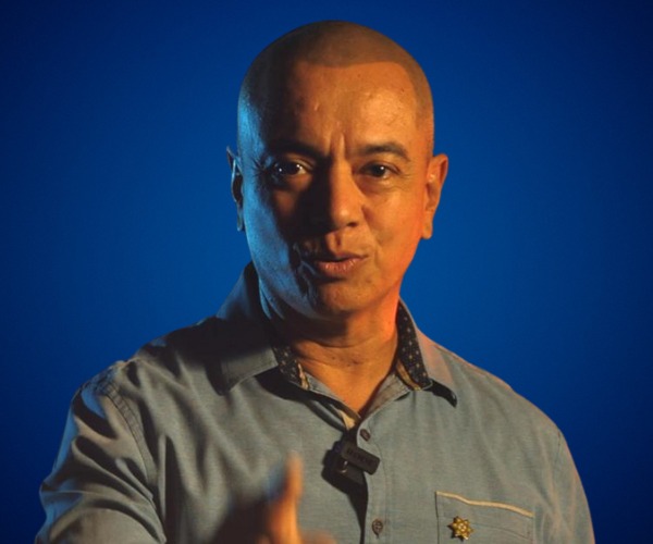 Fotografía de Julian Aguilar Estrada, con expresión decidida, vistiendo camisa azul y micrófono de solapa, sobre un fondo azul intenso con iluminación dramática.