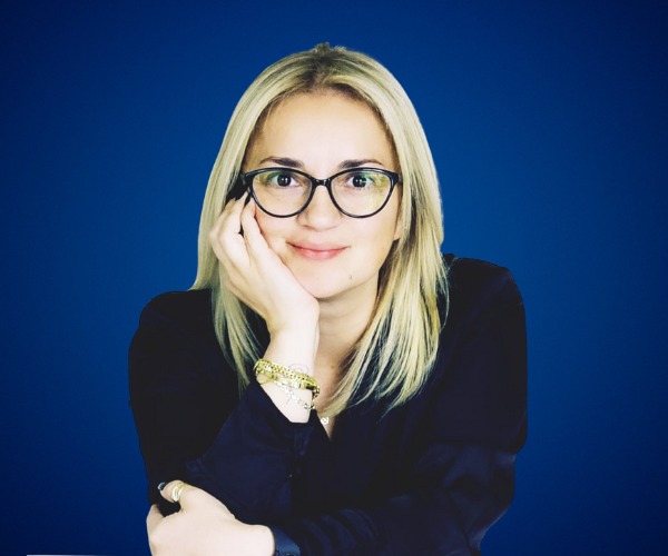 Fotografía de Nancy Reeves, mujer rubia con gafas negras, sonriendo levemente con la mano apoyada en su rostro, sobre fondo azul sólido.