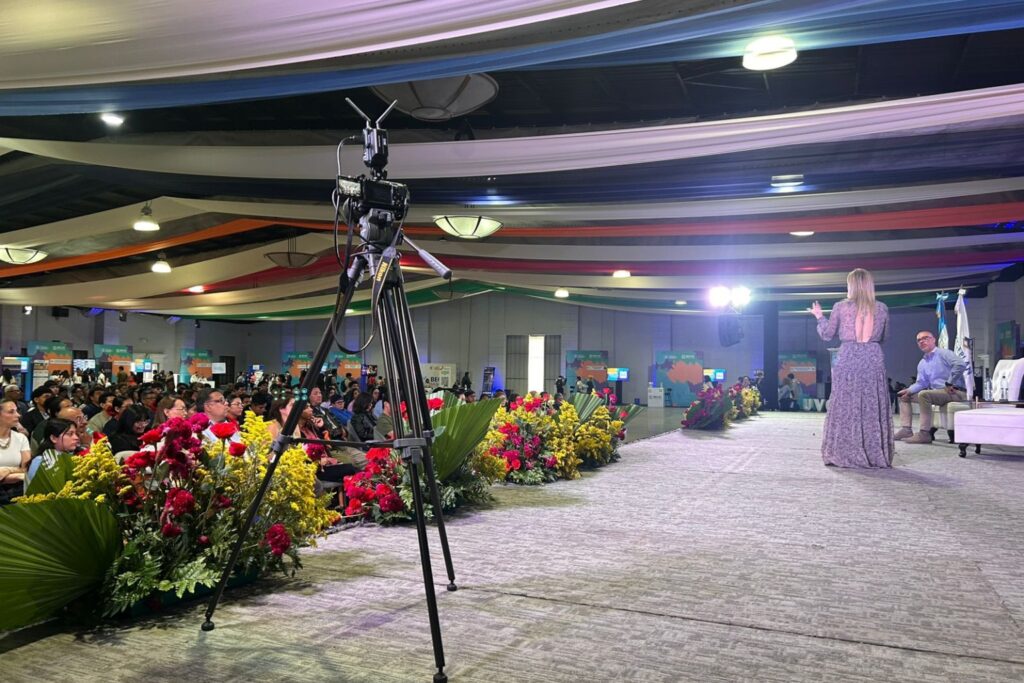Fotografía de Nancy Reeves vista desde el escenario mientras ofrece una charla, vestida elegantemente, frente a un auditorio lleno con decoración floral y techos cubiertos por telas de colores.