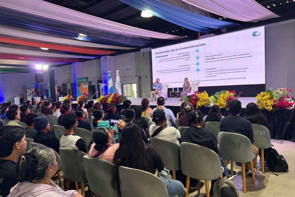 Fotografía de Nancy Reeves sentada en el escenario junto a otro panelista durante una conferencia sobre la evolución de la comunicación humana, con pantalla gigante y público atento.