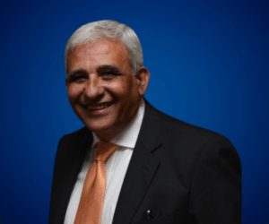 Fotografía de Roberto Patrone, con cabello canoso, sonriendo y vistiendo traje oscuro con corbata naranja, sobre un fondo azul profesional.
