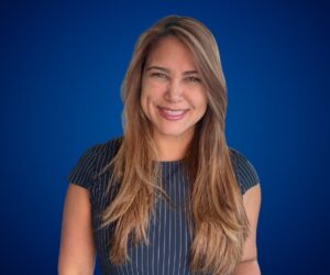 Fotografía de Linda De La Torre sonriendo frente a un fondo azul, con vestido oscuro de líneas finas y cabello largo castaño claro, peinada con raya lateral.