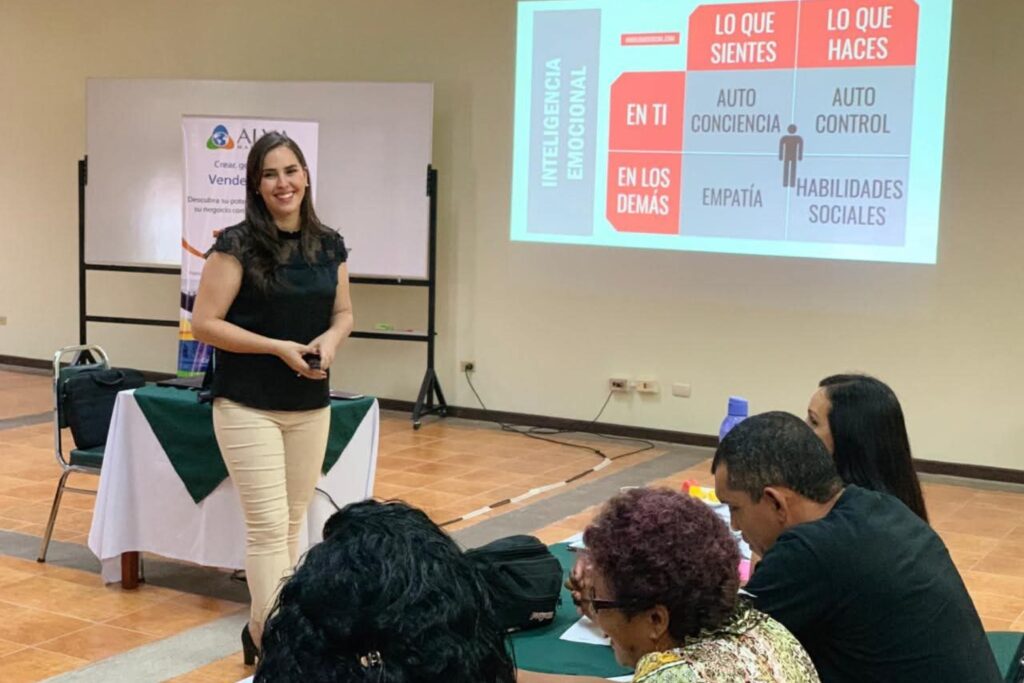 Fotografía de Alejandra Varela Chaverri durante una conferencia sobre inteligencia emocional, de pie frente a una pantalla proyectada, sonriendo mientras interactúa con el público en un salón iluminado y profesional.