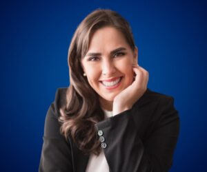 Fotografía de Alejandra Varela, sonriente y vestida con blazer negro, posando con la mano en el rostro frente a un fondo azul liso.