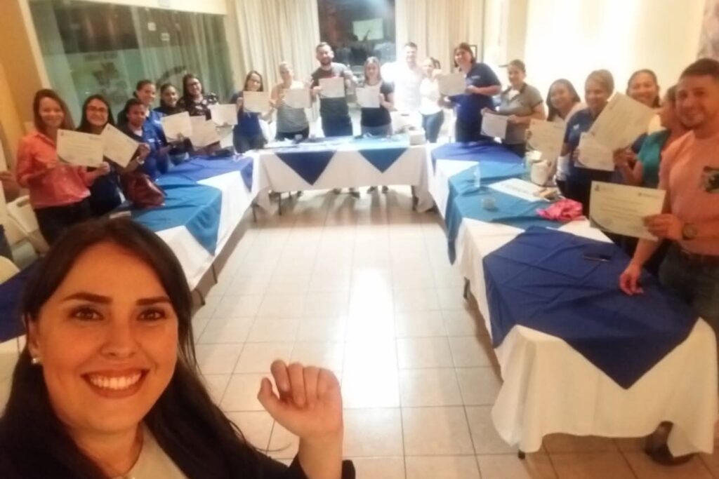Fotografía de Alejandra Varela Chaverri tomando una selfie con un grupo de personas sonrientes que exhiben sus certificados de participación en un salón iluminado con manteles azules.