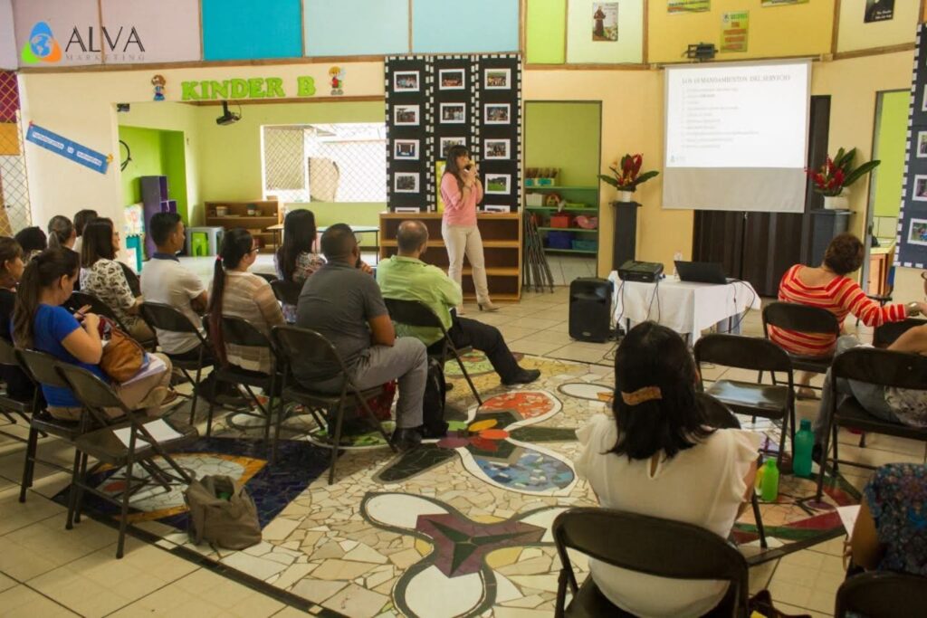 Fotografía de Alejandra Varela Chaverri impartiendo una charla frente a un grupo de adultos en un aula colorida con decoración infantil, mientras proyecta una presentación sobre servicio.
