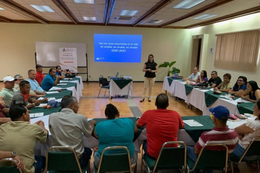 Fotografía de Alejandra Varela Chaverri impartiendo una charla frente a un grupo diverso de personas sentadas en forma de U en un salón de conferencias, con una proyección que muestra una frase inspiradora.
