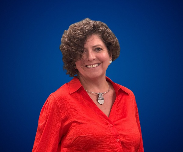 Fotografía de Jennyfer Salvo posando sonriente frente a un fondo azul liso, vestida con una blusa roja y un collar llamativo, en una imagen tipo retrato profesional.