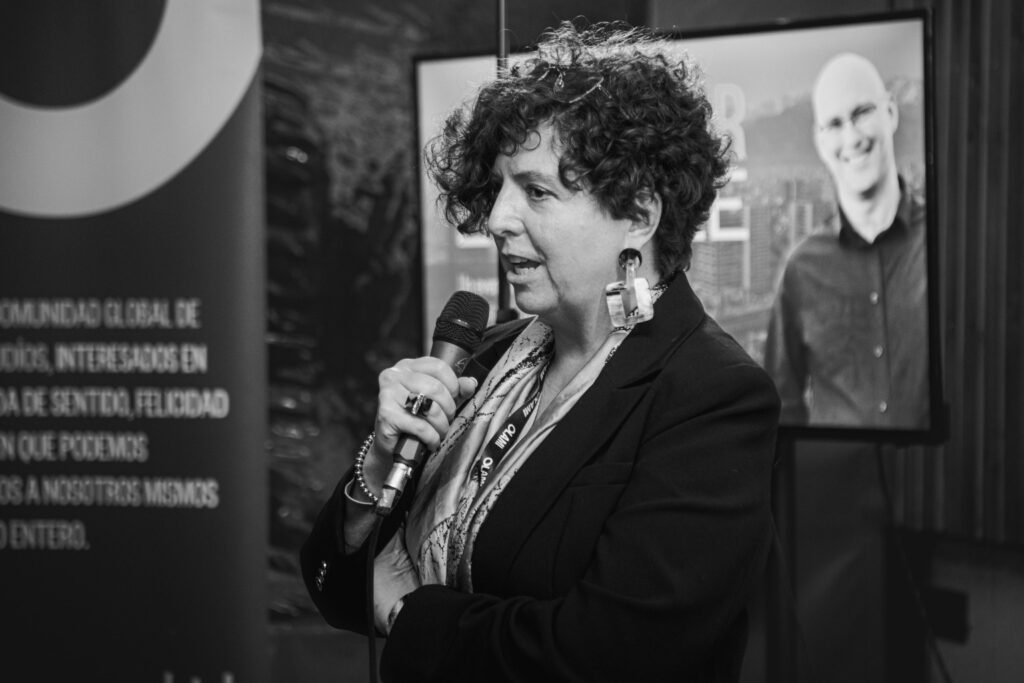 Fotografía en blanco y negro de Jennyfer Salvo hablando con un micrófono en mano durante una conferencia, con expresión concentrada y fondo corporativo desenfocado.